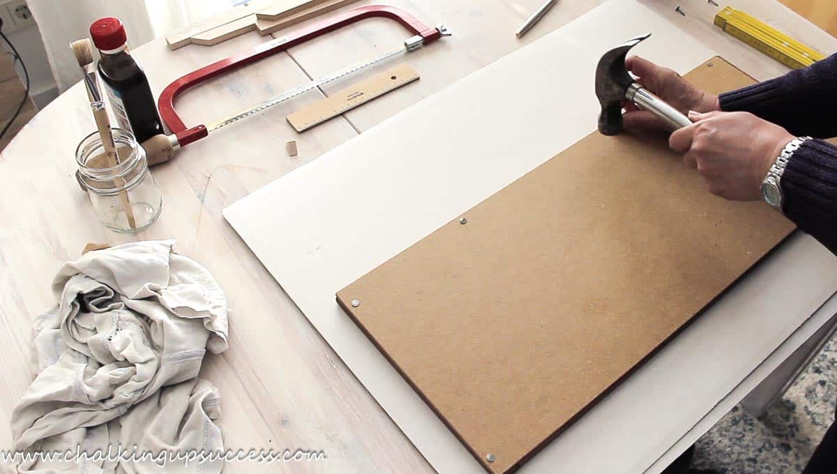 Using a hammer and picture nails to attach a wooden frame to a DIY farmhouse sign.
