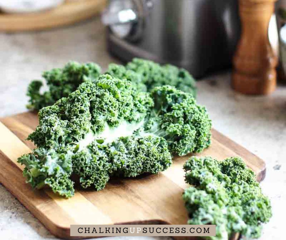 Get the Kale chips recipe on my blog at Chalking Up Success dot com A bunch of curly kale leaves on a wooden chopping board.