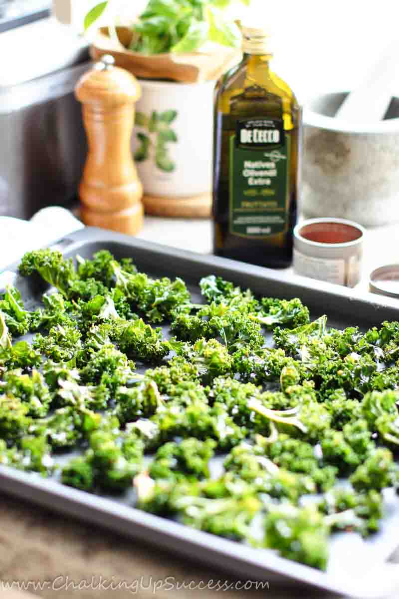 Get the Kale chips recipe on my blog at Chalking Up Success dot com Bite-sized kale chips are spread out on an oven tray before baking.